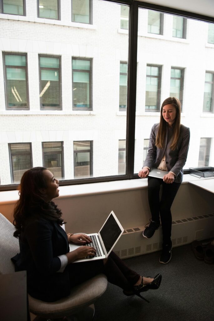 Two businesswomen discuss work in a contemporary office setting with laptops.